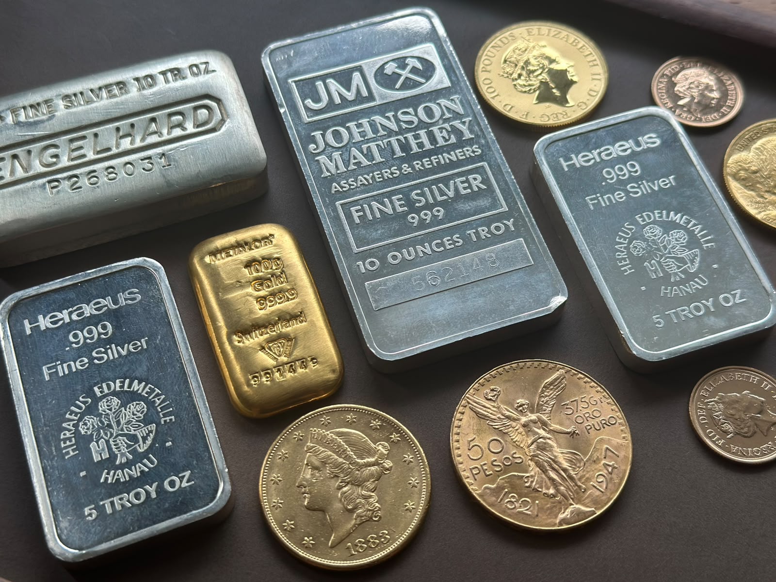 Engelhard, Johnson Matthey and Heraeus silver bars, a Metalor gold bar, and gold coins arranged on a dark surface